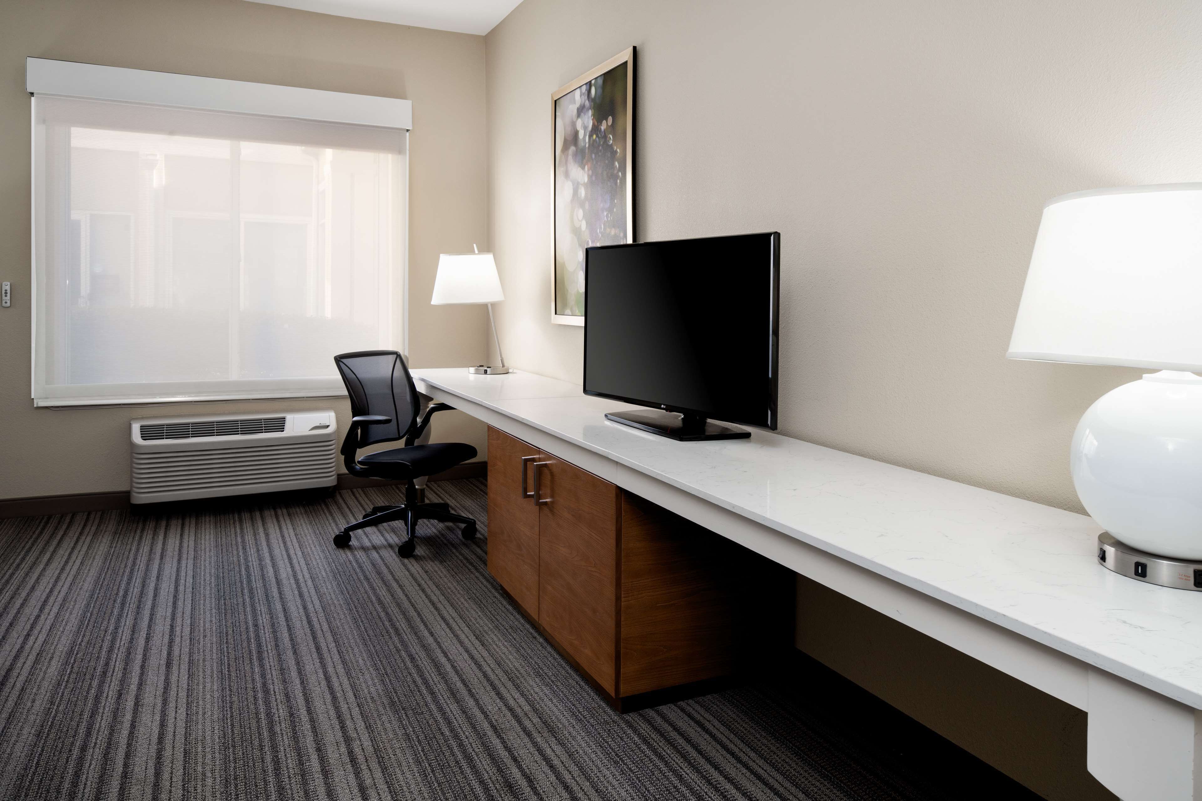 A hotel room with a large window, a flat-screen TV, a desk with a lamp, a chair, and an air conditioning unit.