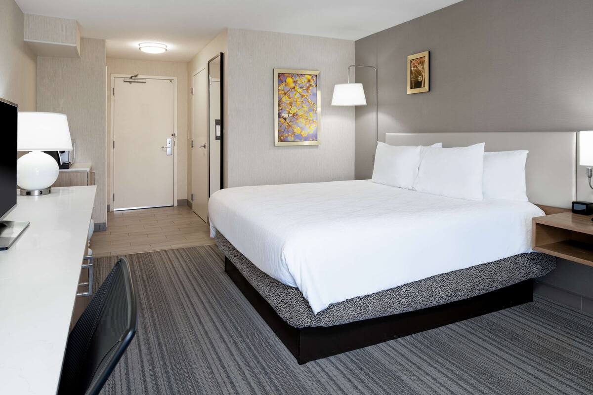 A hotel room with a large bed, a desk with a lamp, a television, and a painting on the wall.