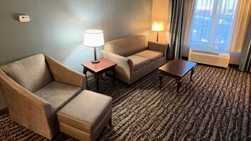 Habitación de hotel con sofá, sillón, reposapiés, una mesita, dos lámparas y un ventanal grande con cortinas.