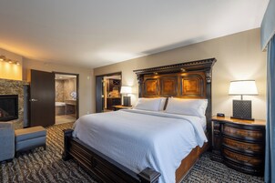 Habitación de hotel con una cama grande, cabecera de madera, mesita de noche con lámpara, cómoda y baño visible a través de una puerta abierta.