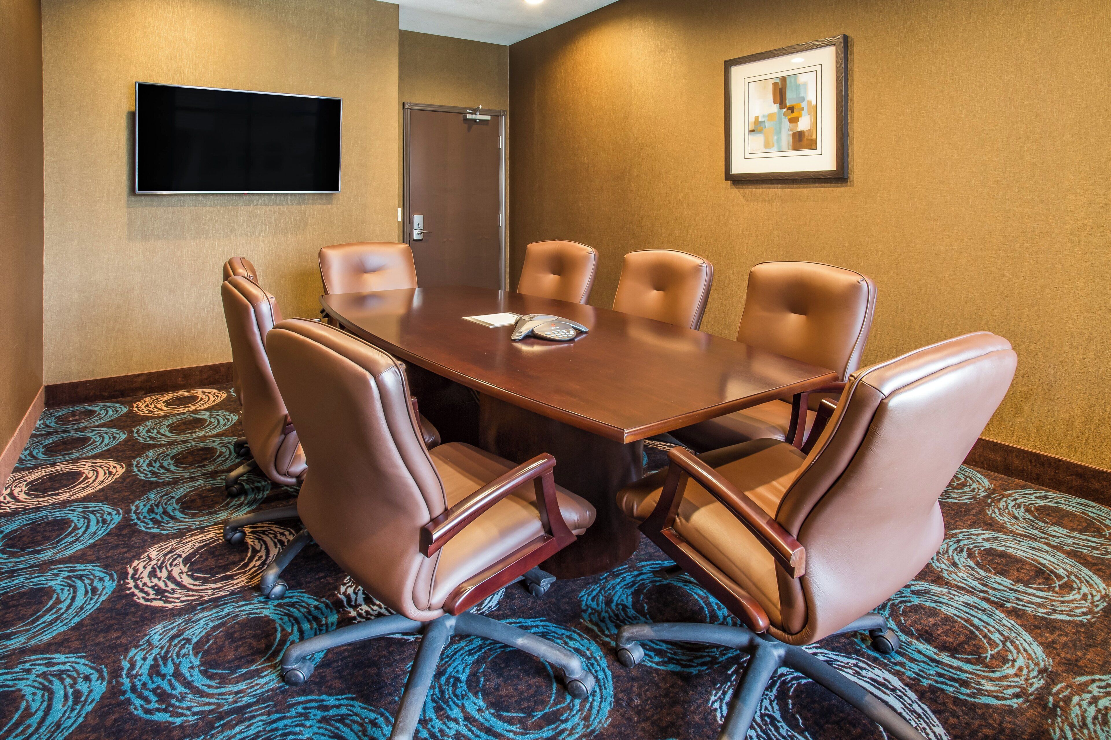 A conference room with a large table, six leather chairs, a flat-screen TV, a door, and a framed picture on the wall.