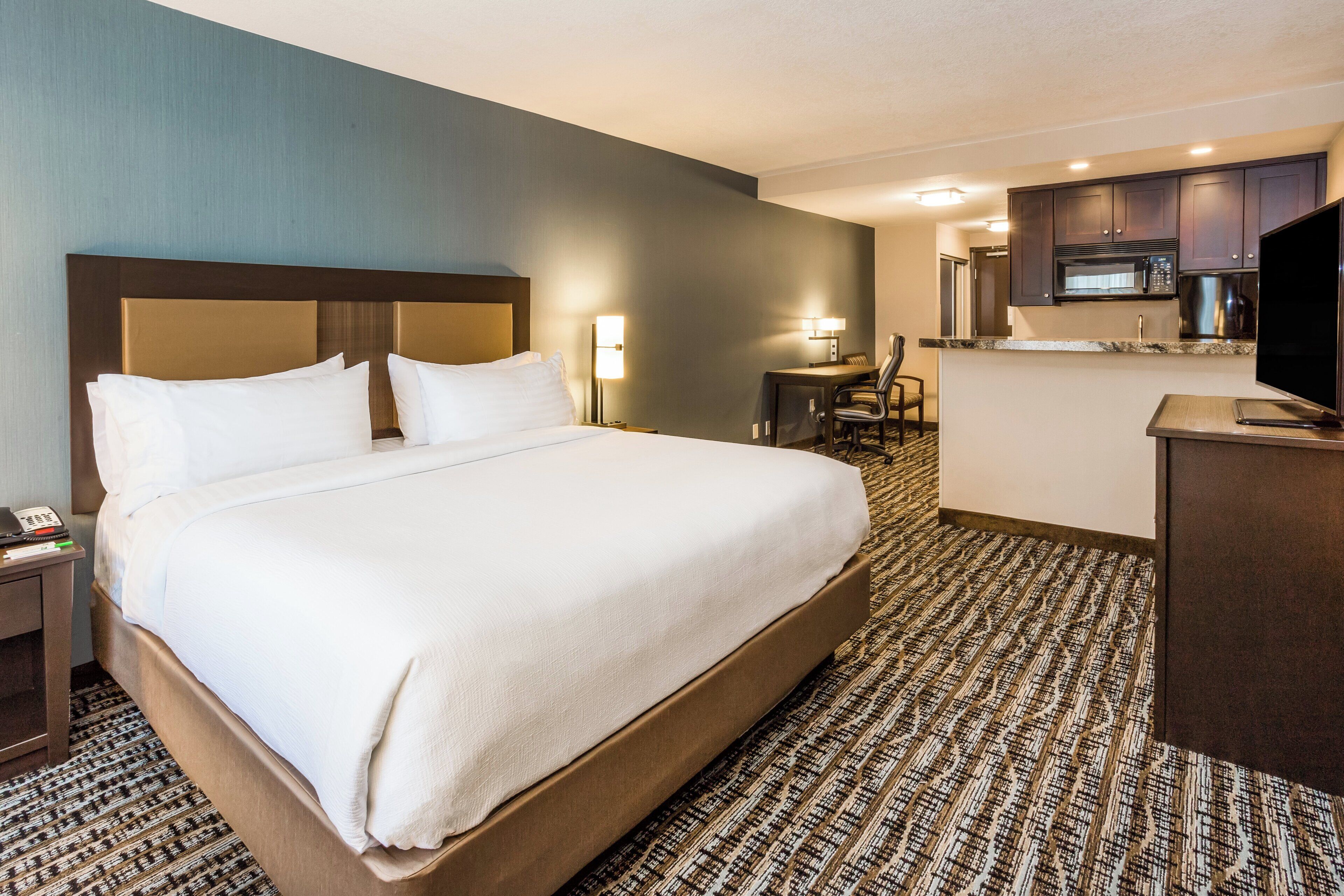 A hotel room with a large bed, a desk, a chair, a television, and a kitchenette.