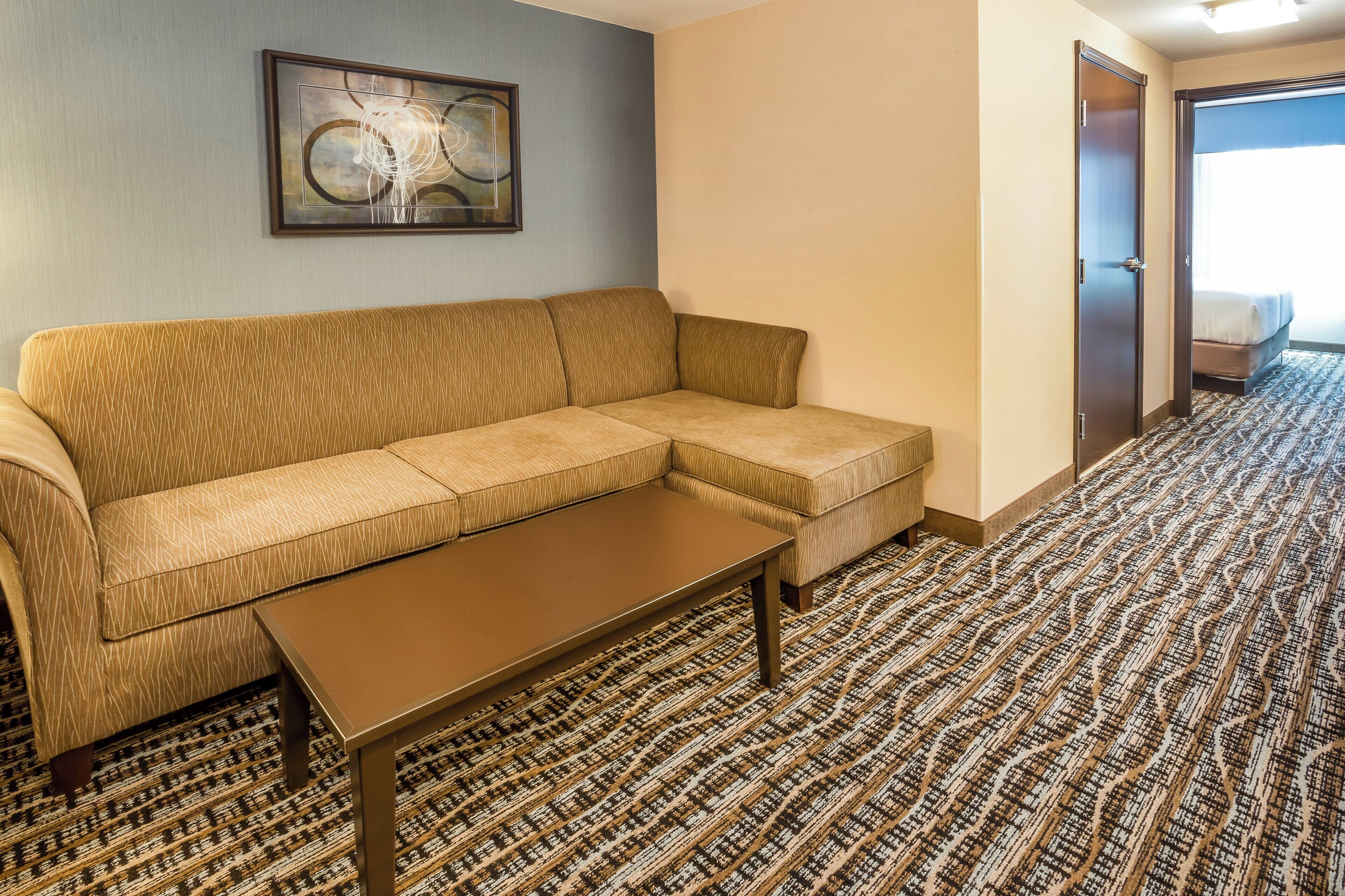 Une chambre d’hôtel avec un canapé, une petite table, une moquette à motifs et une porte donnant sur une chambre à coucher.