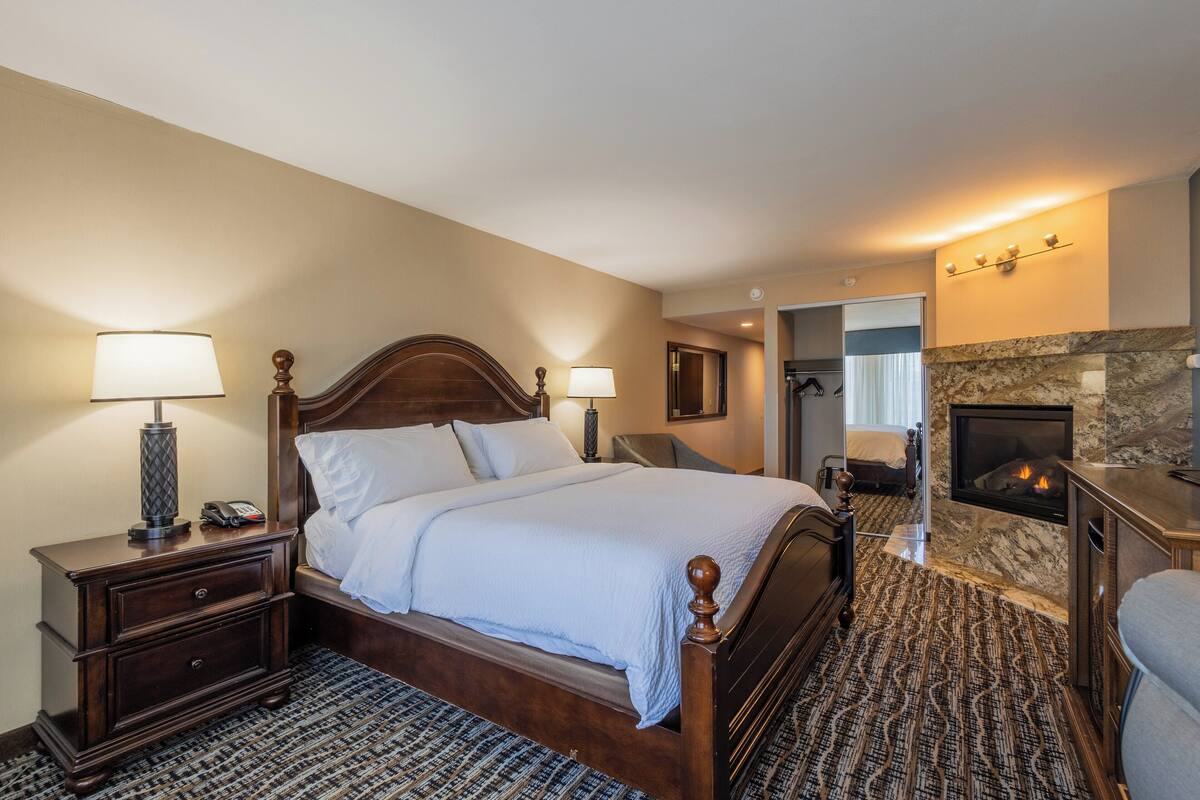 A hotel room with a large bed, a nightstand with a lamp, a fireplace, and a seating area with a chair.