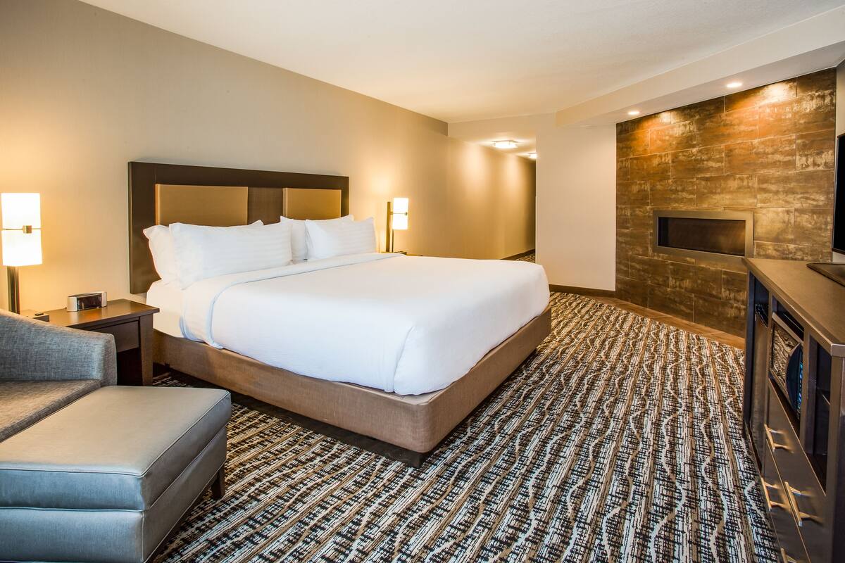 A hotel room with a large bed, a flat-screen TV, a fireplace, and a patterned carpet.