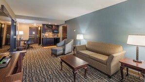 A hotel room with a living area featuring a sofa, armchairs, a coffee table, and a desk with a computer. There is a kitchen area with a microwave and a refrigerator.