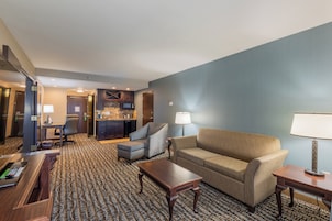 Habitación de hotel con sala de estar que incluye un sofá, sillones, una mesa de centro y un escritorio con una computadora. Hay una zona de cocina con microondas y refrigerador.