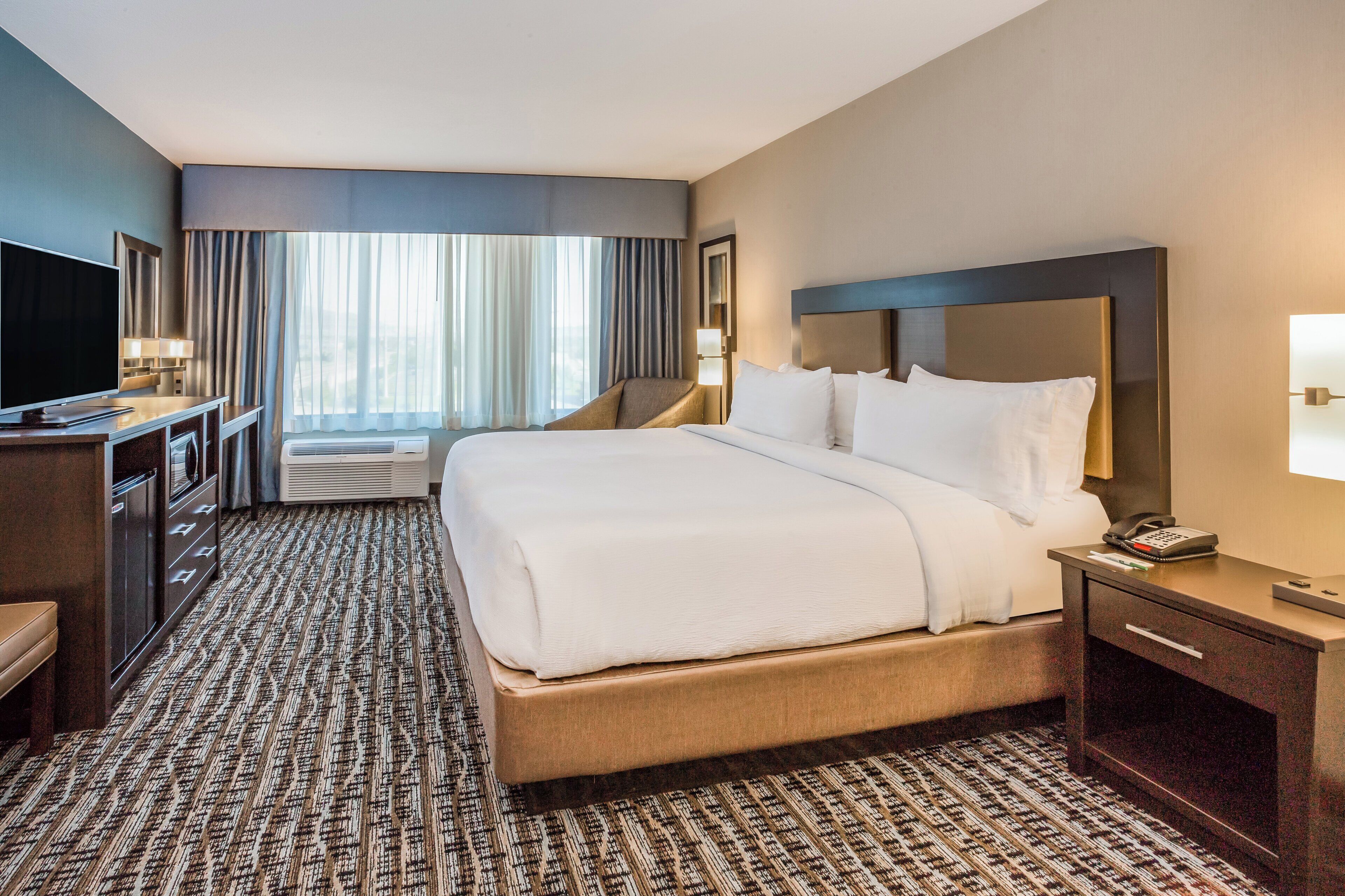 A hotel room with a large bed, a desk with a flat-screen TV, a bedside table with a lamp, and a large window with curtains.