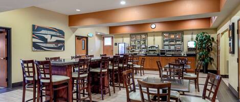 A hotel lobby with multiple dining tables and chairs, a kitchen area with a counter and appliances, and a large abstract painting on the wall.