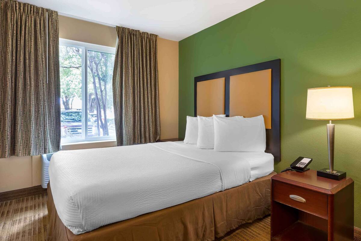 Habitación de hotel con una cama grande, una mesita de noche con lámpara y una ventana con cortinas.