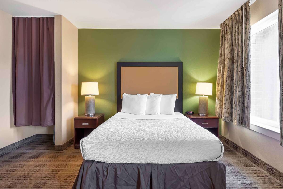 Habitación de hotel con una cama grande, dos mesitas de noche con lámparas, paredes verdes y una ventana con cortinas.