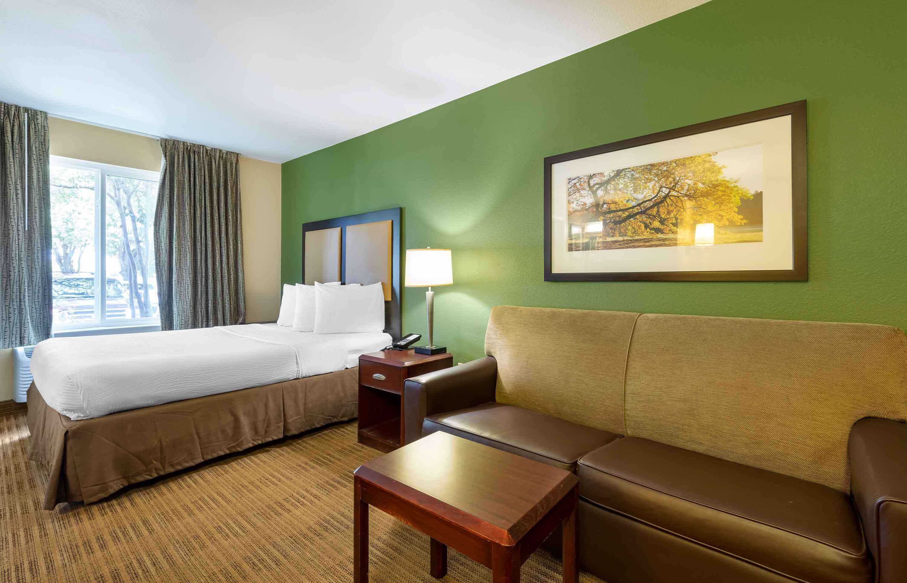 A hotel room with a bed, a sofa, a small table, a nightstand, a lamp, and a framed picture on the wall.