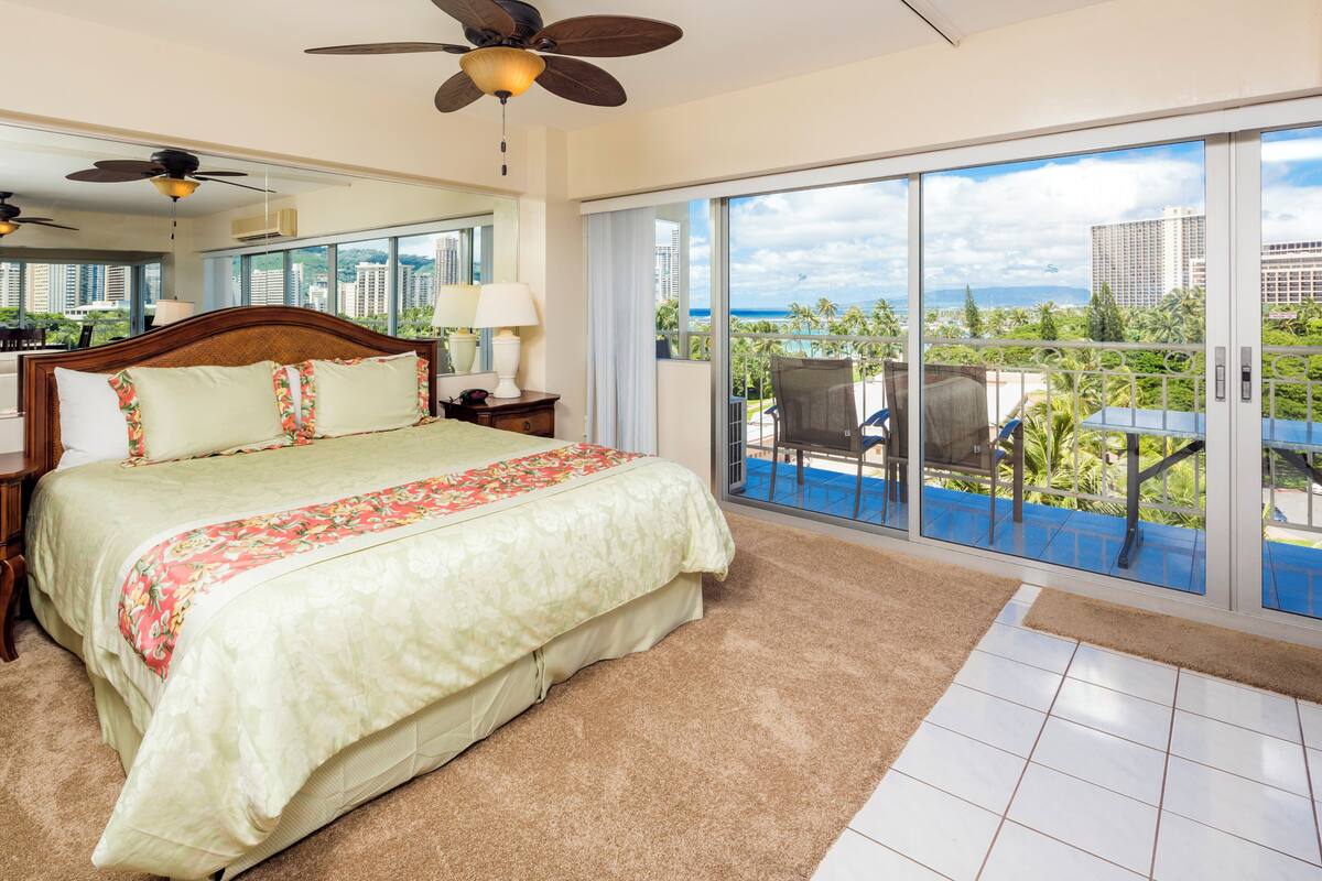 Quarto de hotel com uma cama grande, ventilador de teto, vista para o oceano e varanda com mesa e cadeiras.