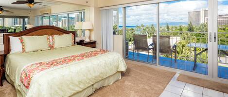 A hotel room with a large bed, a ceiling fan, a view of the ocean, and a balcony with a table and chairs.
