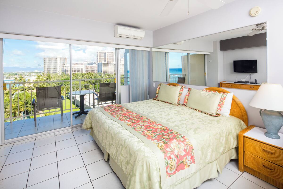 Quarto de hotel com varanda, uma cama com roupa de cama florida, uma televisão e uma mesa de cabeceira de madeira.