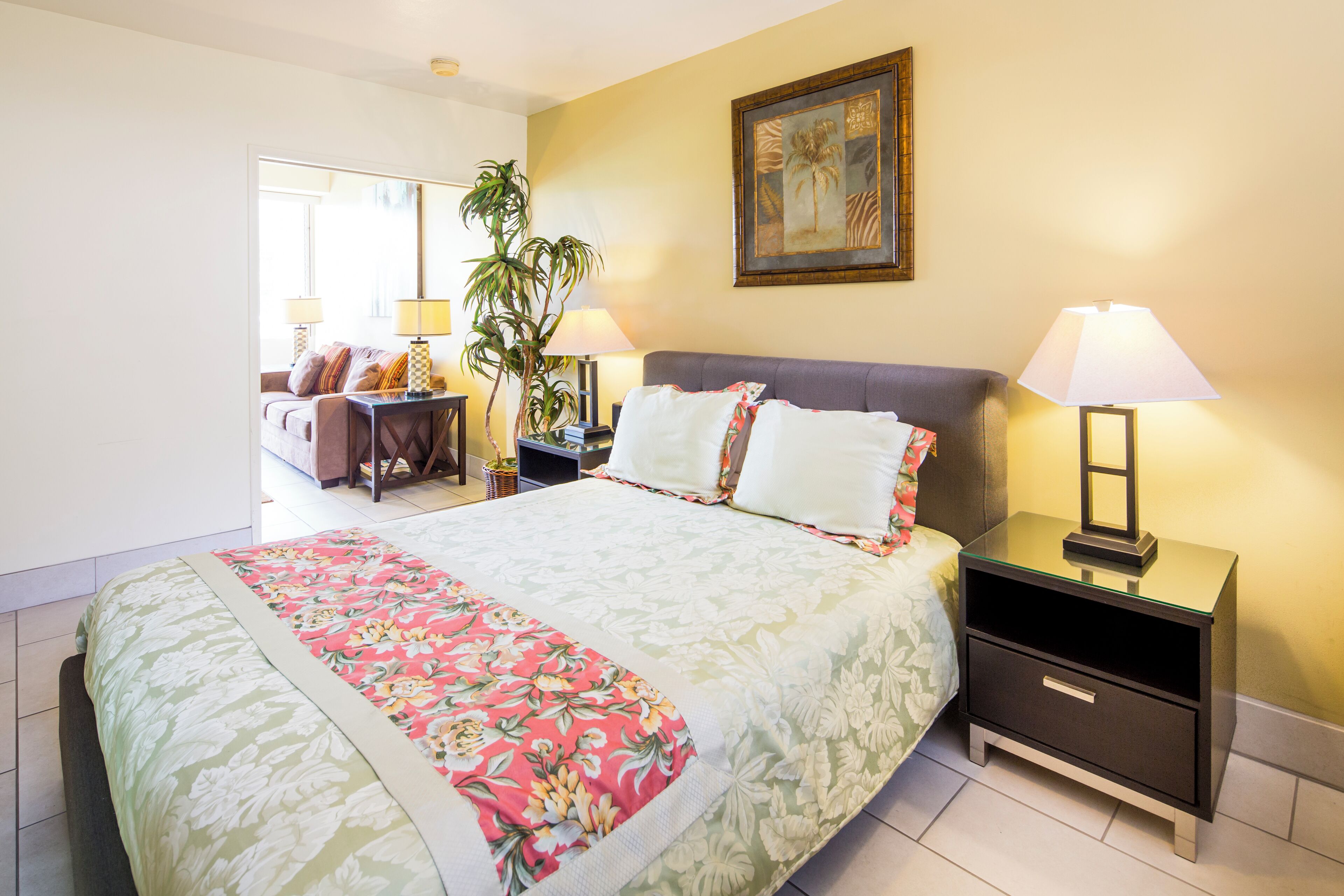 A bedroom with a bed, nightstands, a lamp, a plant, and a painting on the wall.