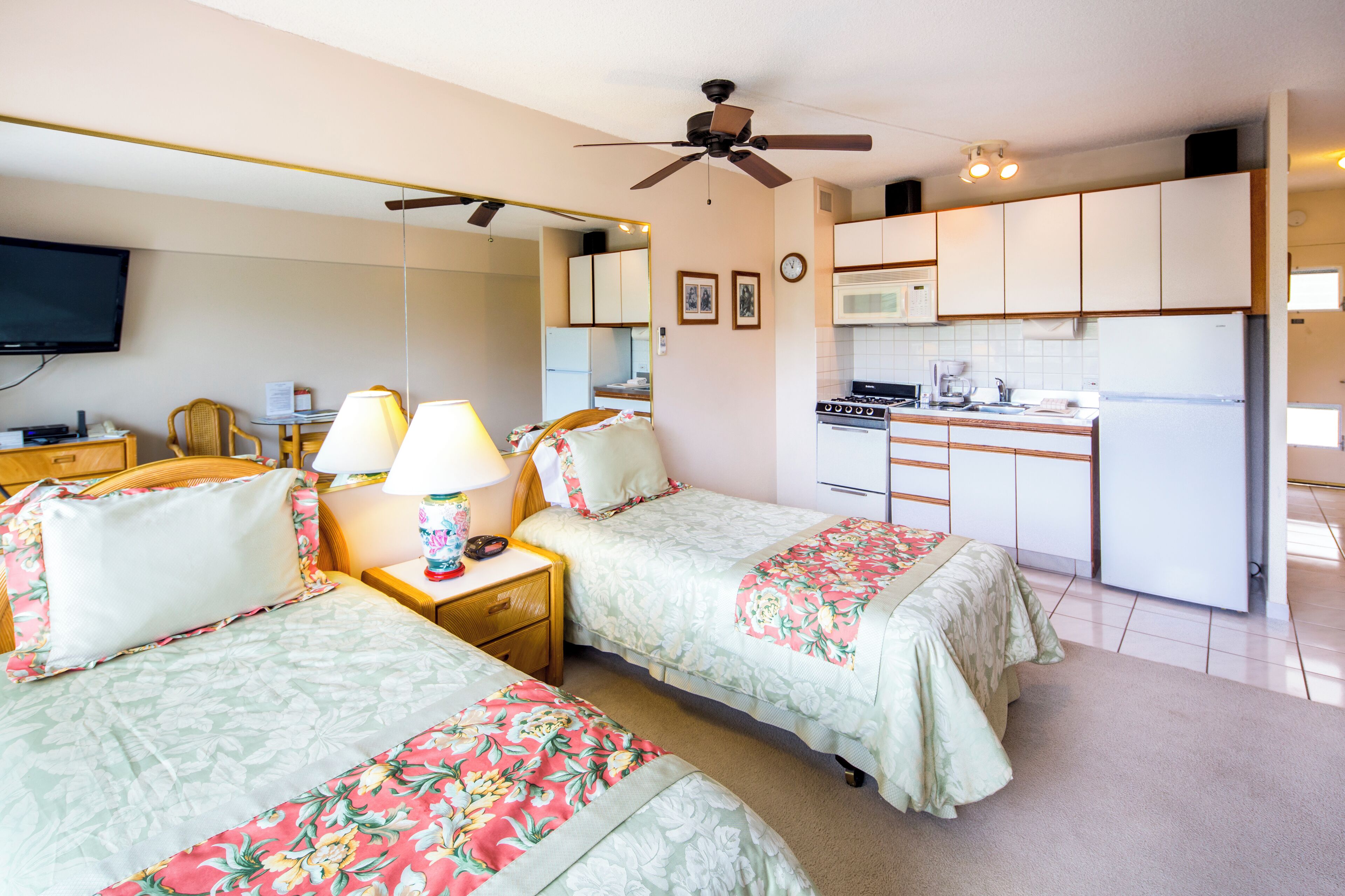 Una habitación de hotel compacta con dos camas, una kitchenette, un ventilador de techo, un televisor de pantalla plana y una pequeña mesa con lámparas.
