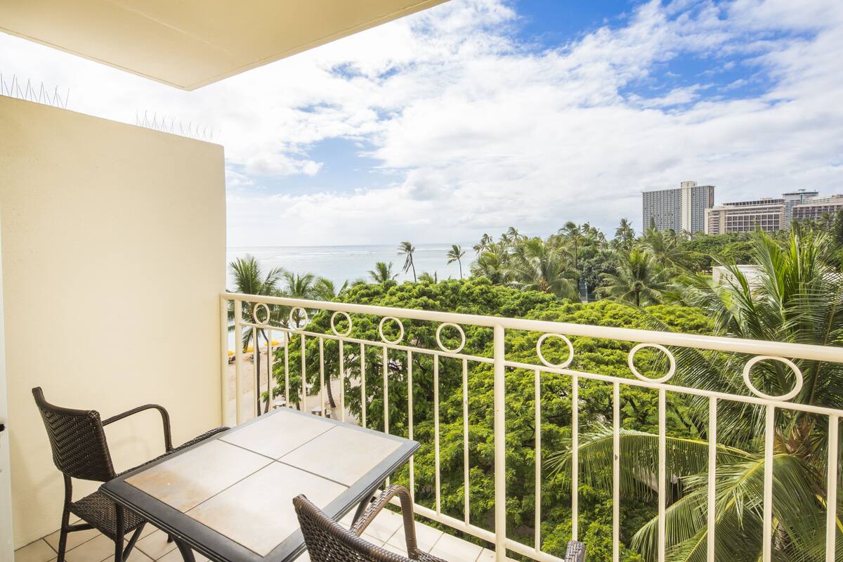 Varanda com mesa e cadeiras, vista para um jardim exuberante e um horizonte urbano com edifícios altos.