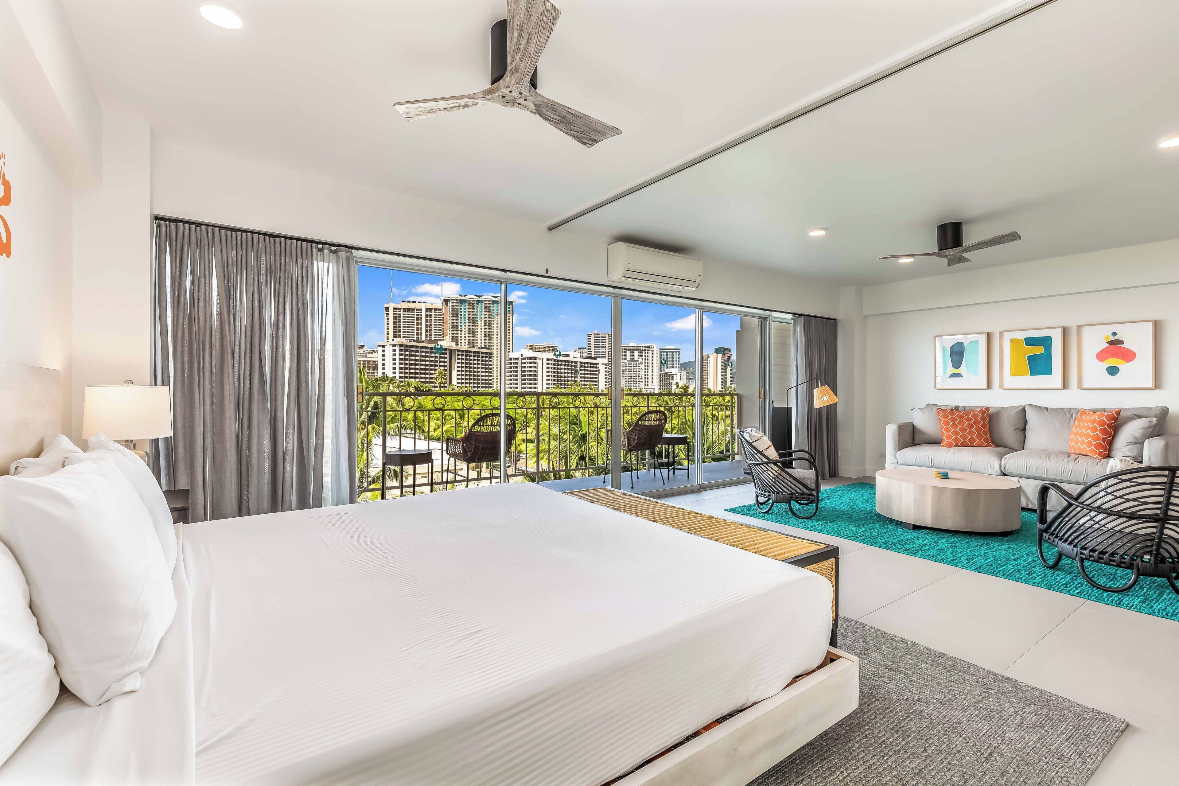 A modern hotel room with a large bed, a balcony with outdoor furniture, a sofa, and a coffee table.