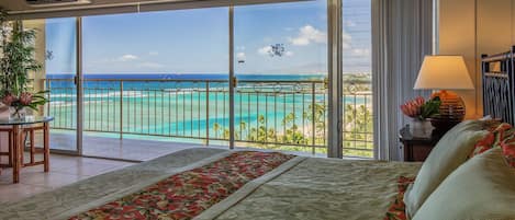 Un cuarto de hotel con una cama grande, un balcón con vista al océano y una planta tropical en la mesita de noche.