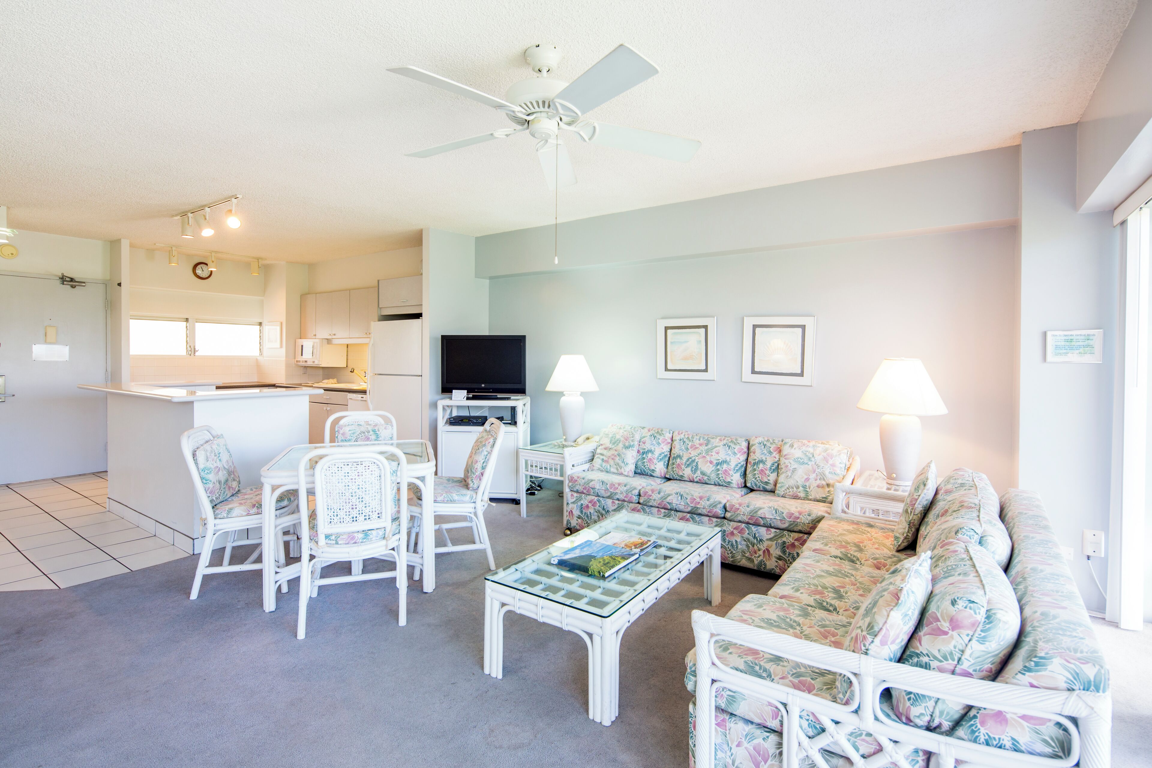 A spacious living room with a white sofa, a glass coffee table, and a dining area with white chairs. A ceiling fan and a kitchen with a TV are visible in the background.