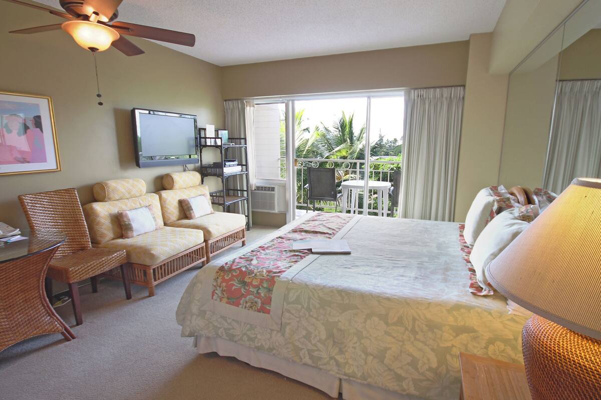 Quarto de hotel com cama, sofá, televisão, varanda com mesa e cadeiras, e ventilador de teto.