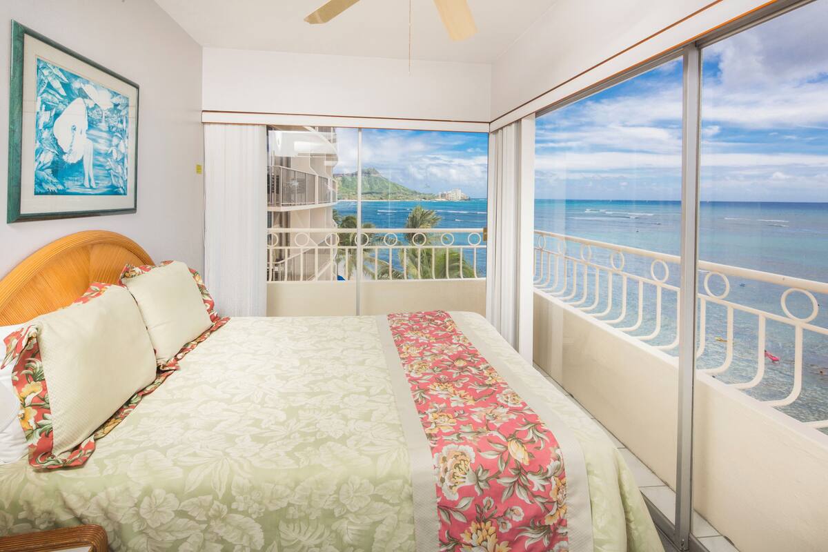 Quarto com cama, uma grande janela com vista para o mar e varanda com grade.