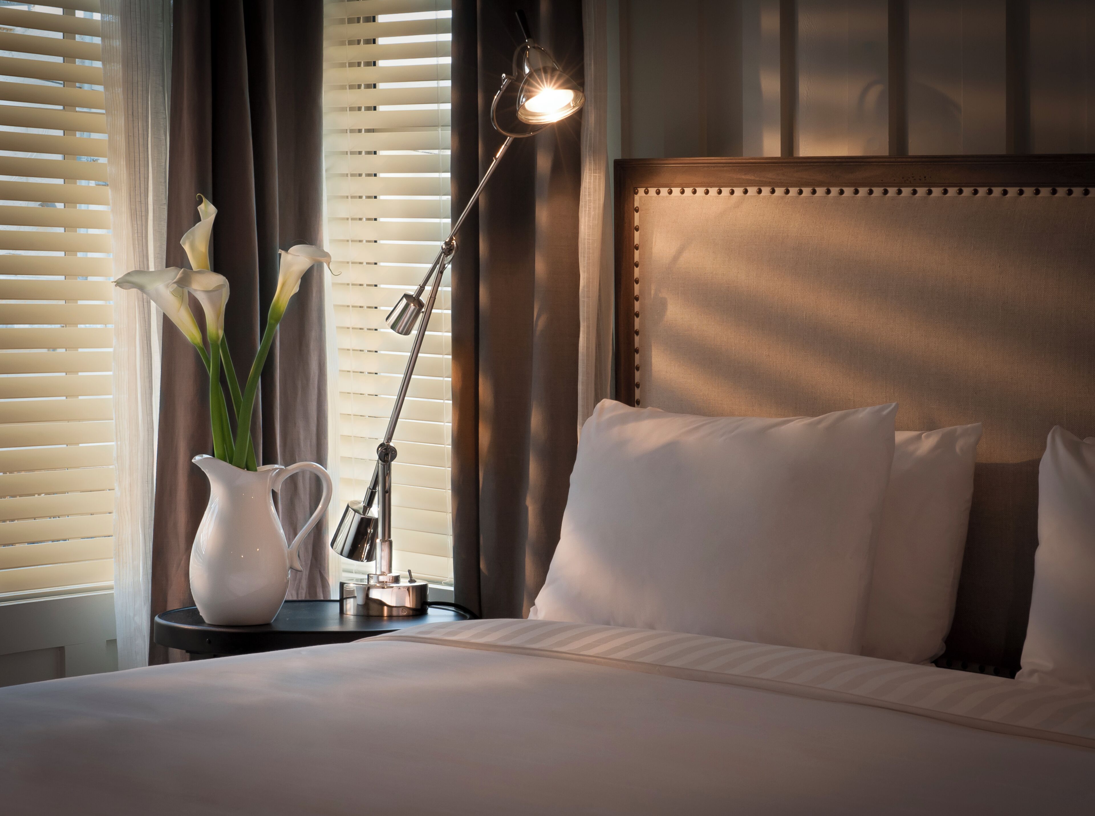 A neatly made bed with white linens, a bedside lamp, and a vase with flowers.