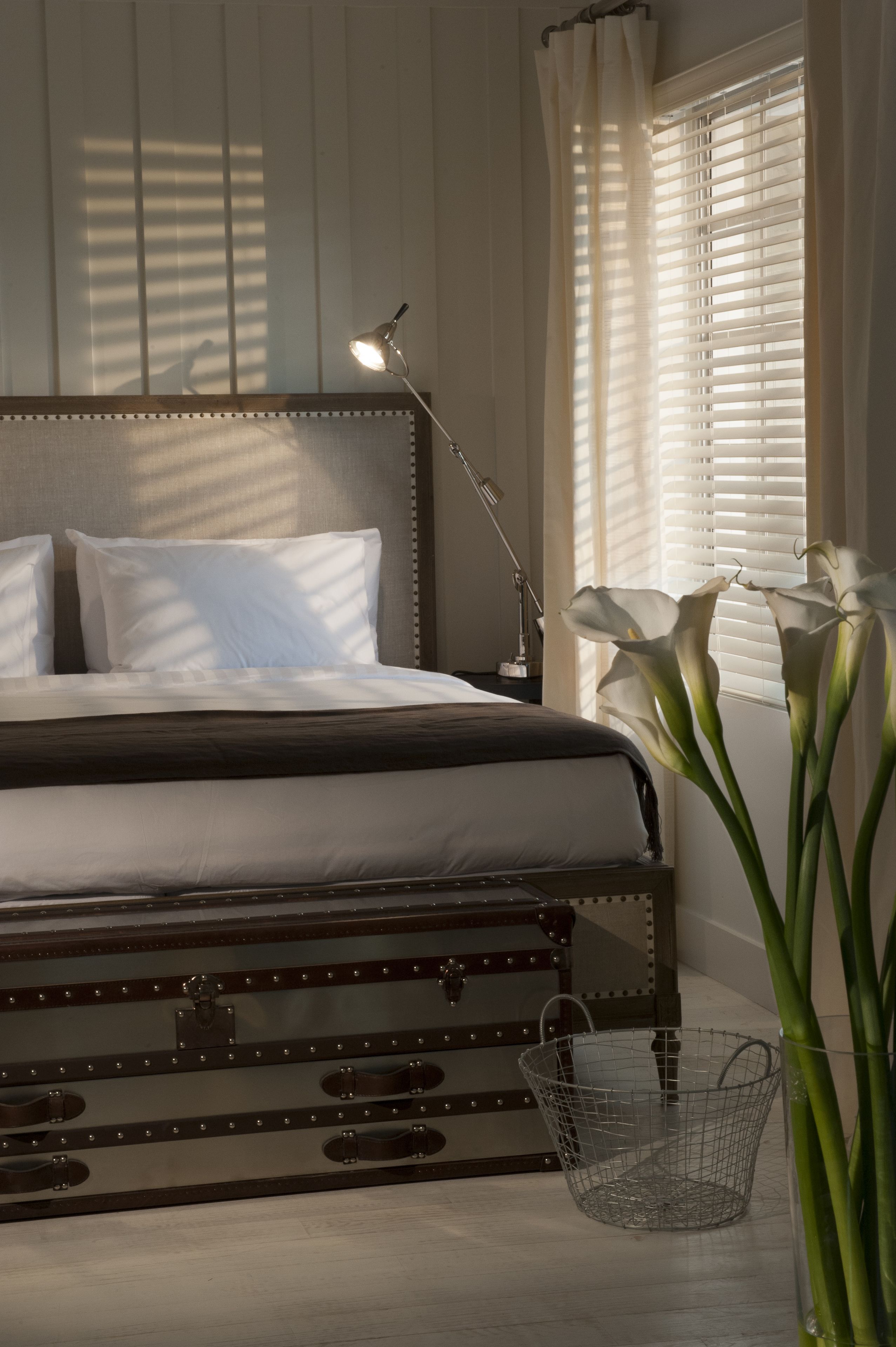 A neatly made bed with white linens, a bedside lamp, and a wire basket on the floor.