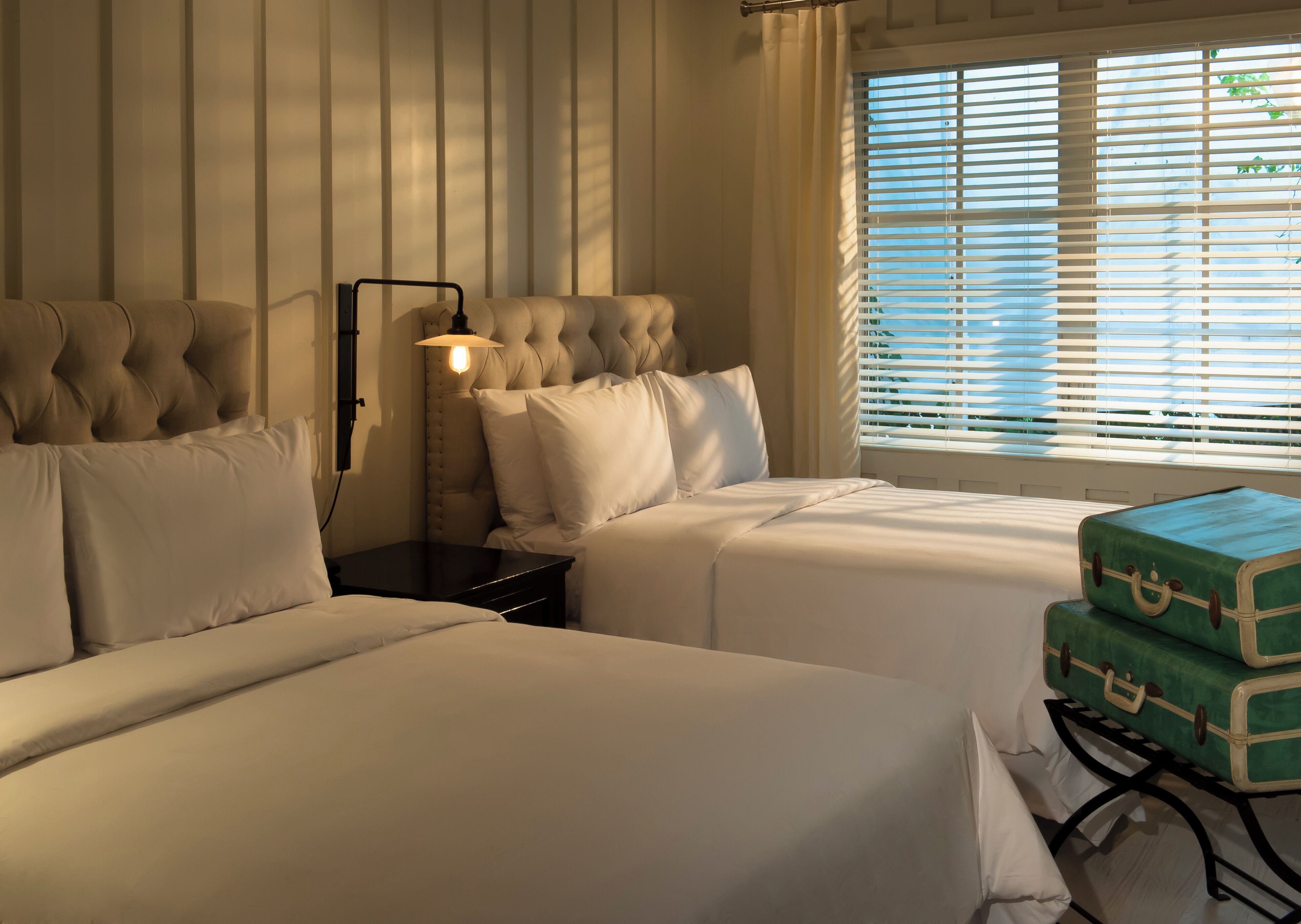 A hotel room with two beds, a nightstand with a lamp, and a window with blinds.