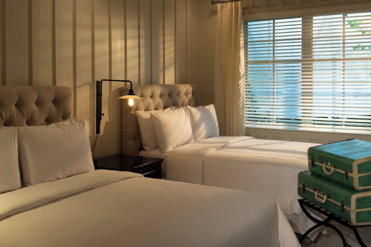 A hotel room with two beds, a nightstand with a lamp, and a window with blinds.