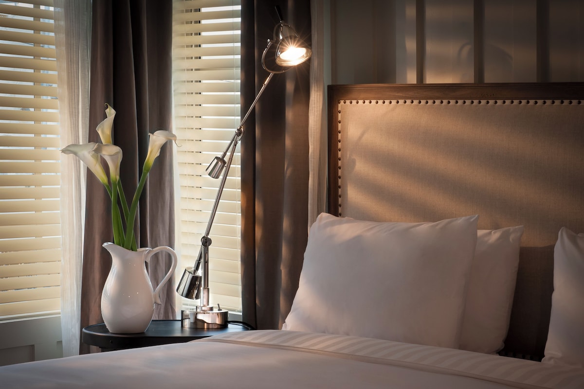 A neatly made bed with white linens, a bedside lamp, and a vase with flowers.