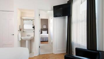 Quarto de hotel com uma cama, uma televisão fixada na parede, um banheiro com pia e espelho, e vista para o corredor.
