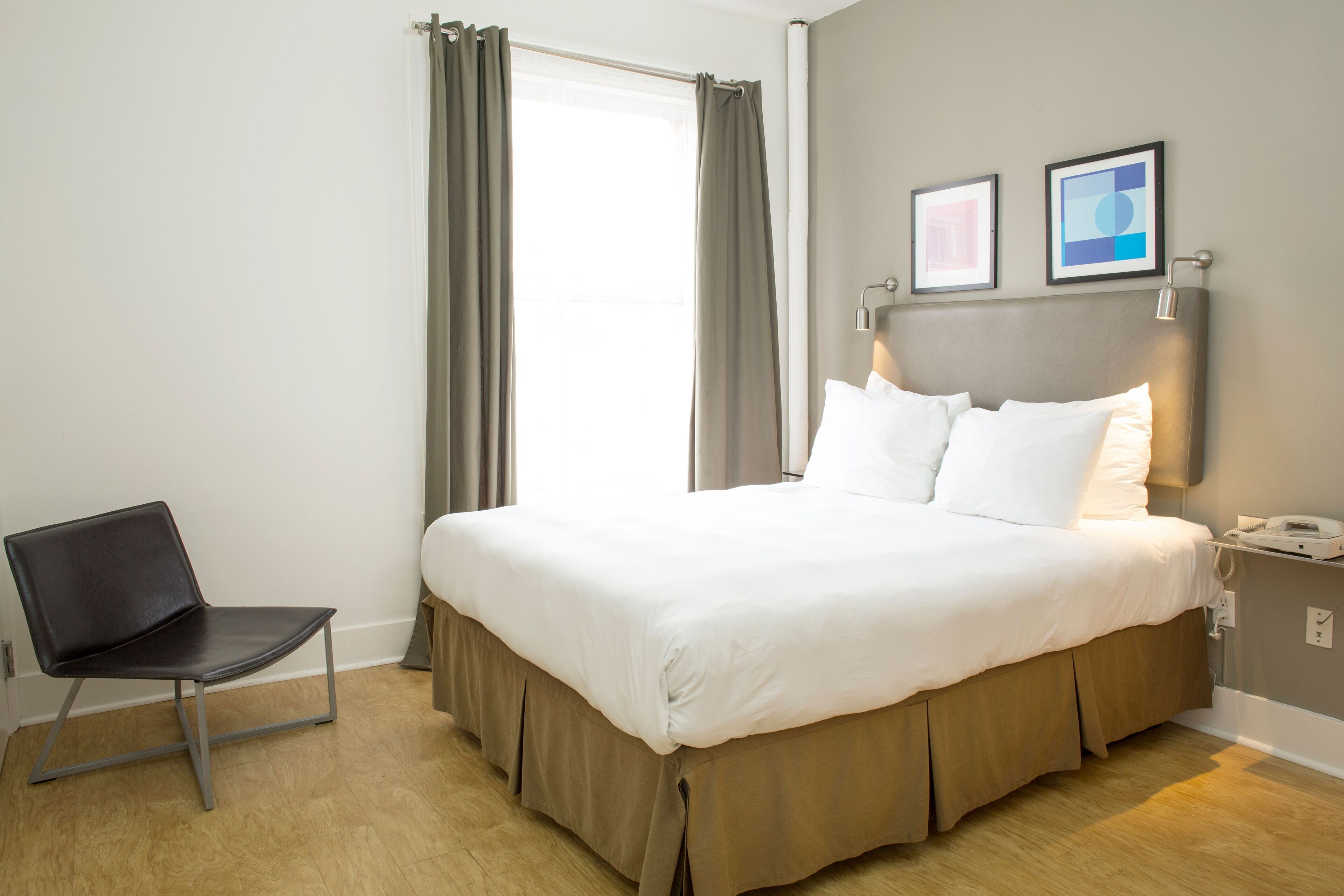 Une chambre d’hôtel bien rangée, avec un lit, une chaise et une table de chevet sur laquelle se trouve une lampe.