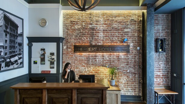 Un espace d’accueil avec un mur en briques, une horloge et une enseigne indiquant « HÔTEL HERBERT ».