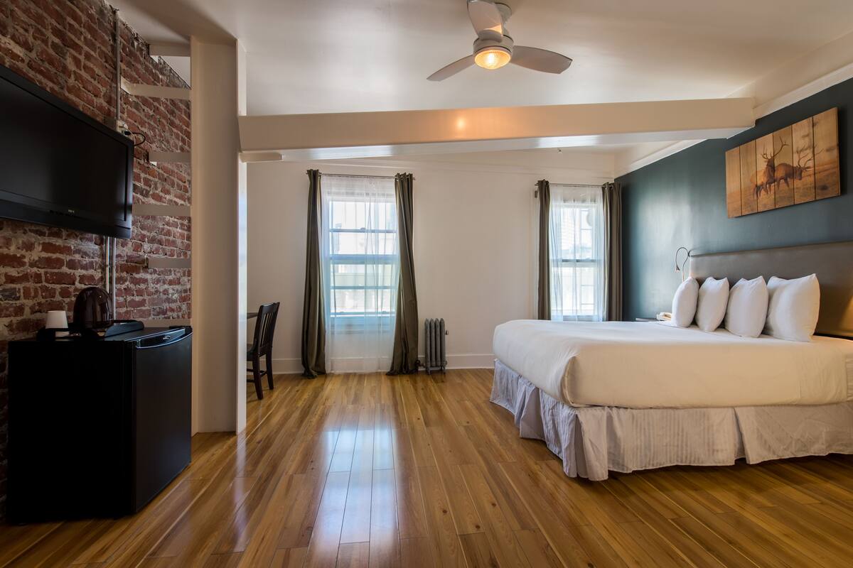 Una habitación de hotel con pared de ladrillo, suelo de madera, cama con ropa blanca, escritorio con lámpara y ventilador de techo.