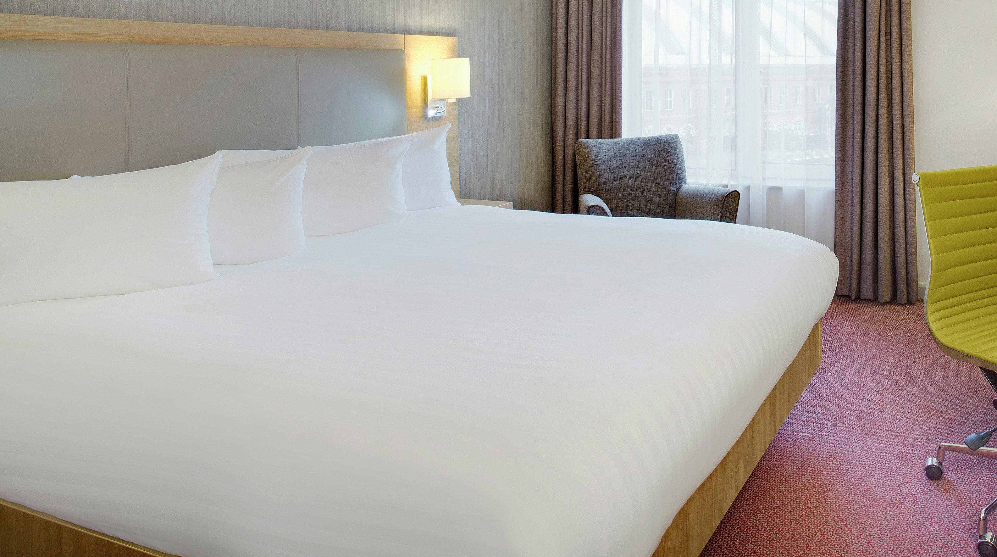 Una cama grande con sábanas blancas, un sillón tapizado gris y una silla amarilla en una habitación de hotel con alfombra roja.