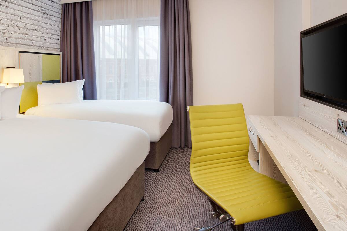 Habitación de hotel con dos camas, un escritorio, una silla, un televisor y una ventana con cortinas.