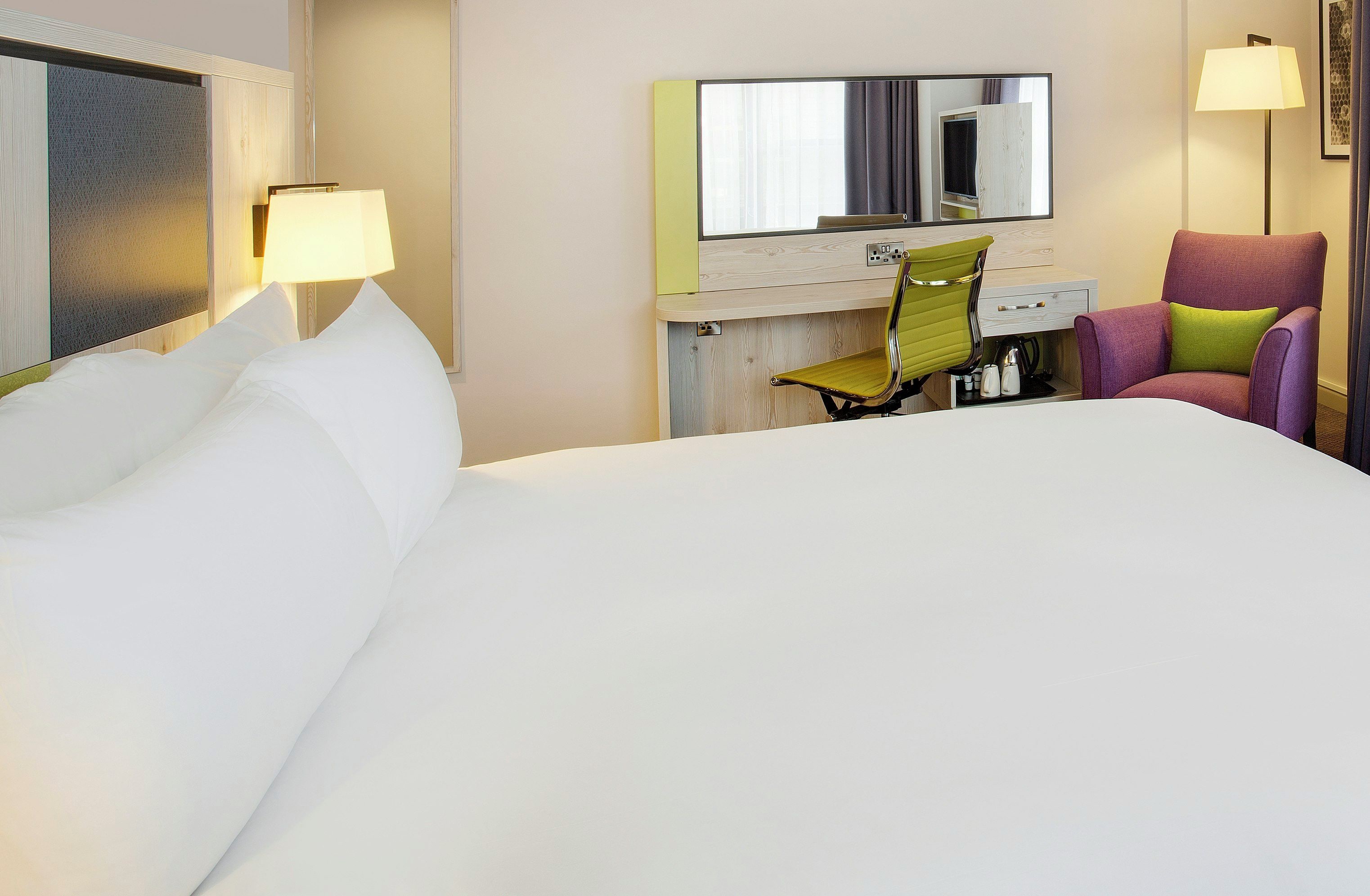 A hotel room with a large bed, a desk with a chair, a mirror, and a lamp.