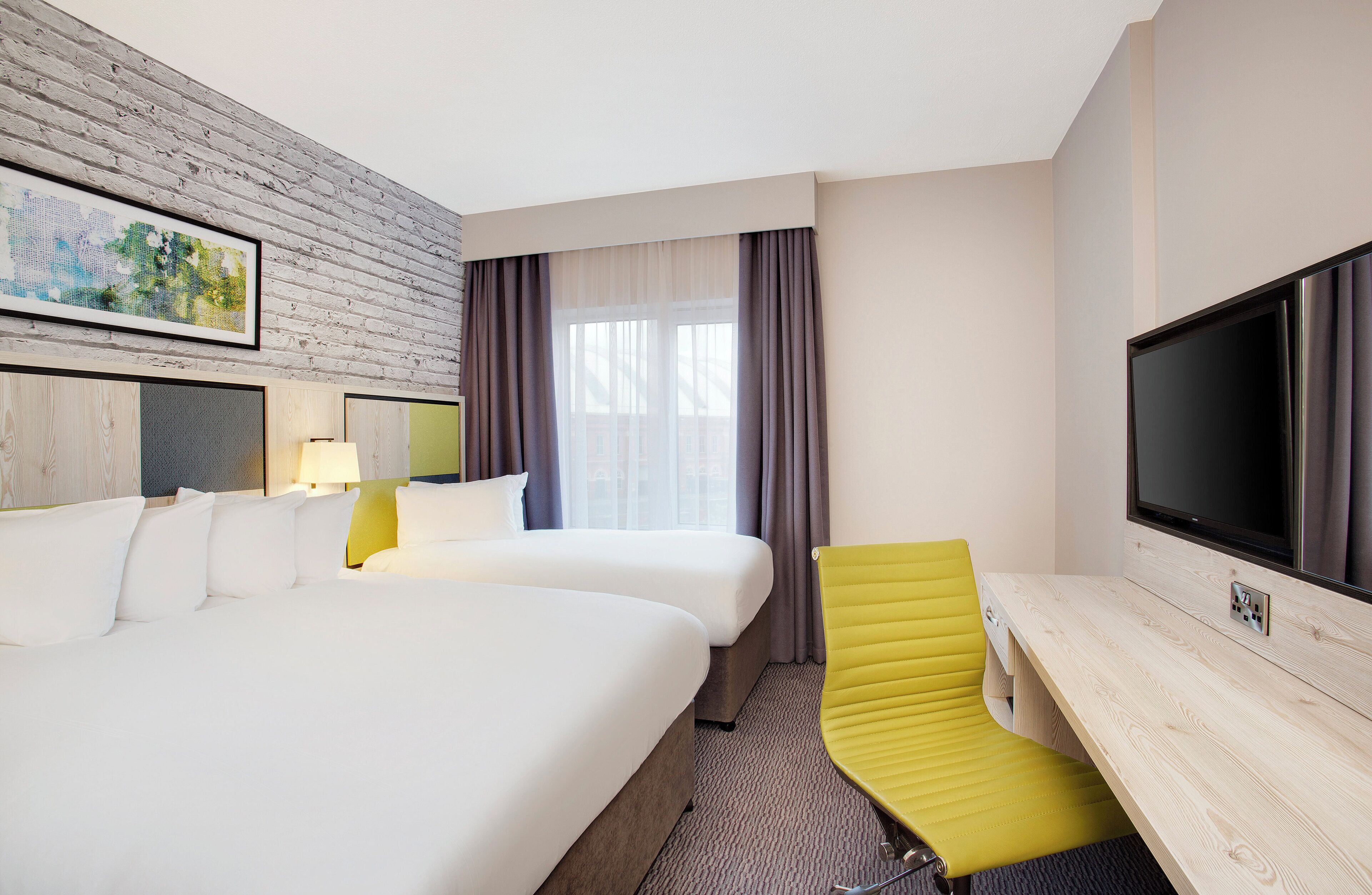 A hotel room with two beds, a desk, a television, and a window with curtains.
