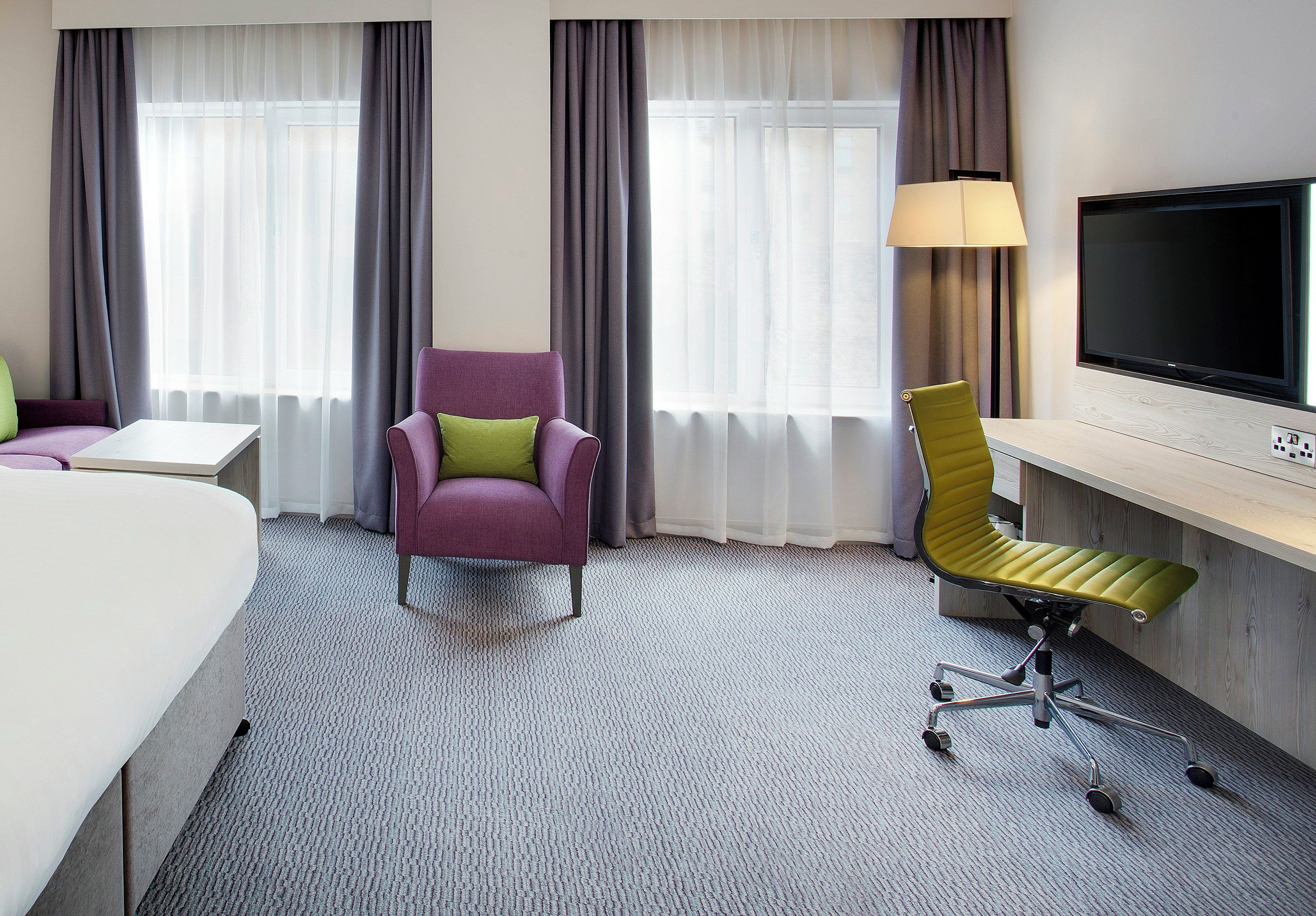 A hotel room with a bed, a desk, a purple armchair, a green office chair, a TV, and two windows with curtains.