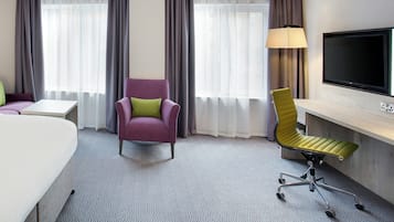 A hotel room with a bed, a desk, a purple armchair, a green office chair, a TV, and two windows with curtains.