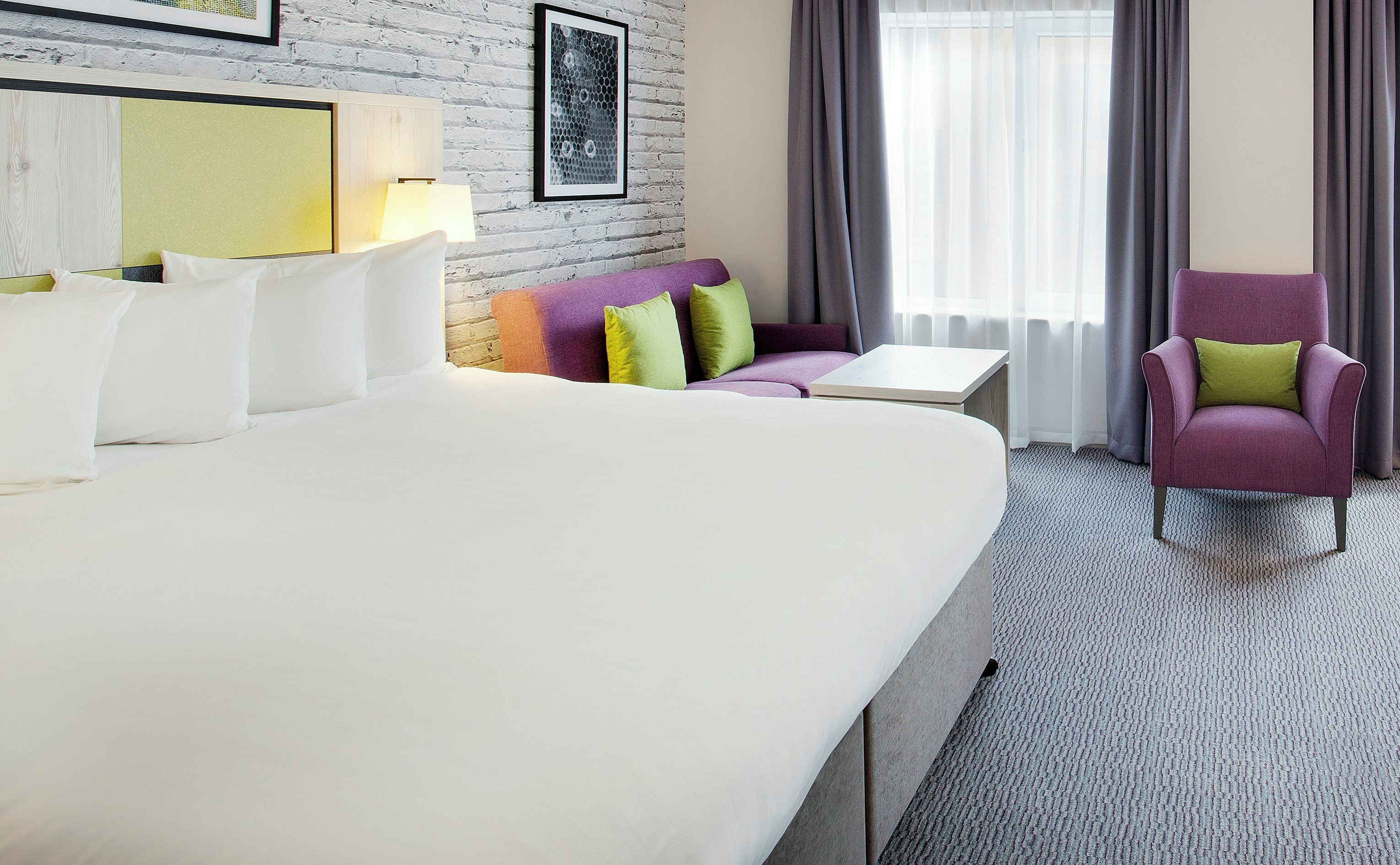A hotel room with a large bed, a small table, a purple armchair, and a sofa with colorful cushions.