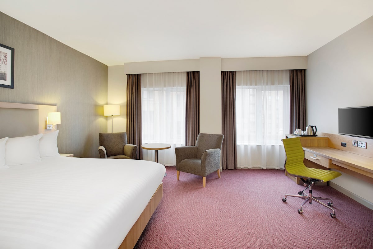 Una habitació d'hotel amb un llit gran, dues butaques, un escriptori amb una cadira, un televisor i una taula petita.