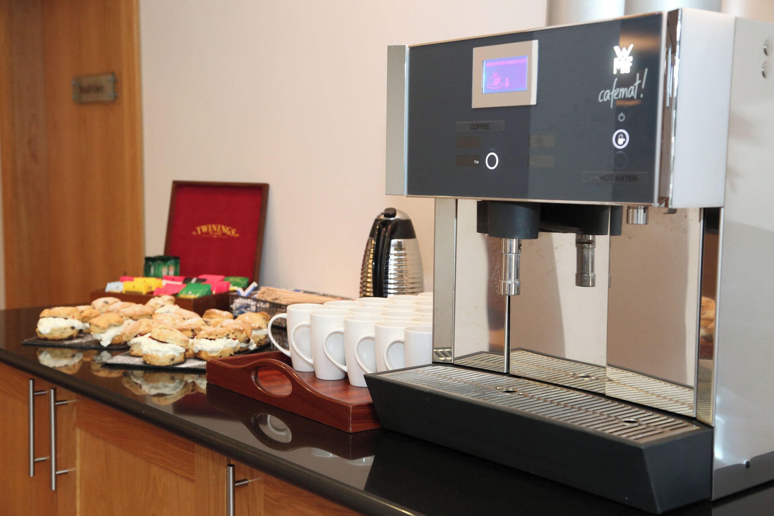 Una cafetera con pantalla digital, una bandeja de pasteles y varias tazas blancas sobre una encimera.