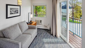 A hotel room with a grey sofa, a framed picture on the wall, a window with a view of greenery and mountains, and a balcony with a railing.