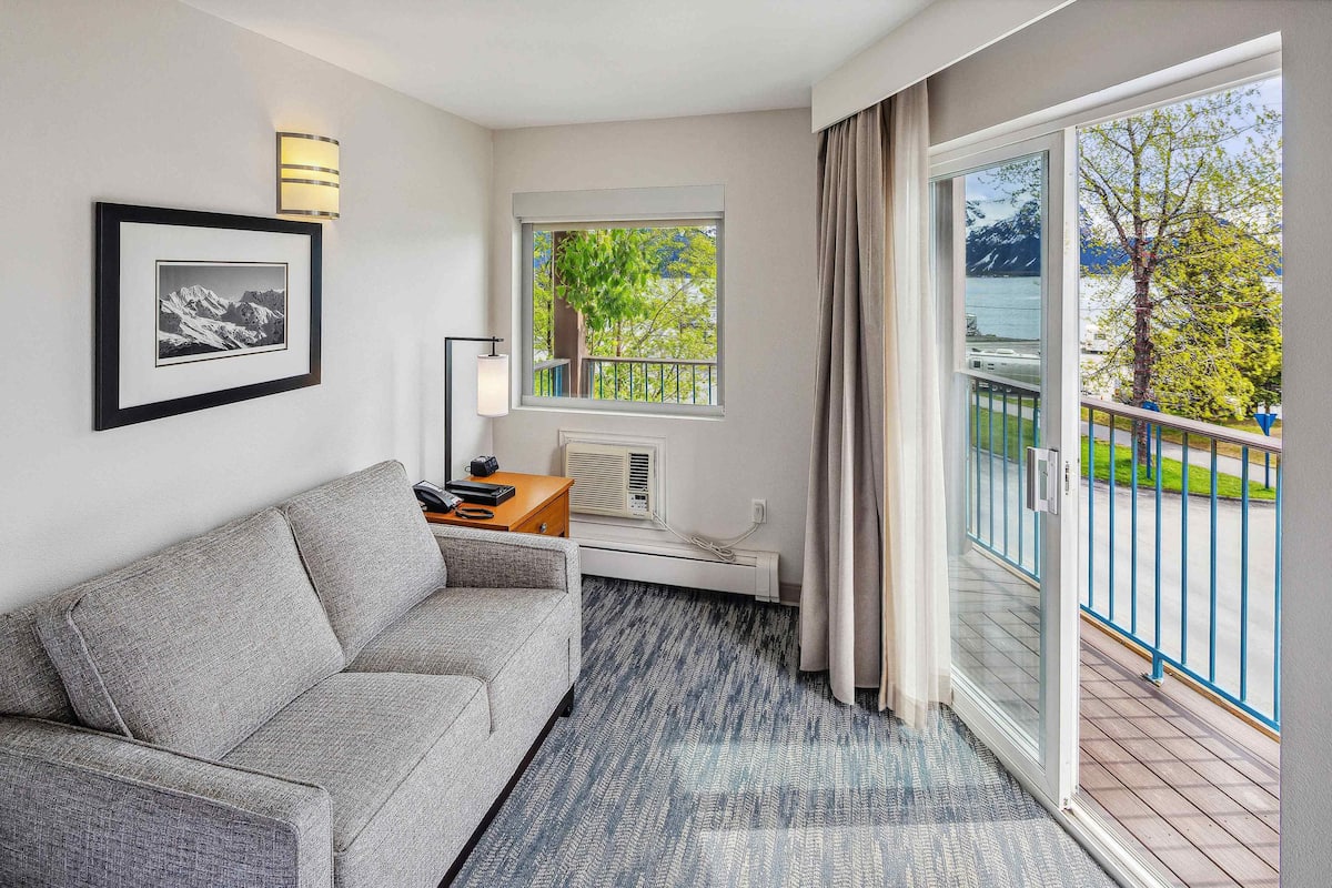 Une chambre d’hôtel avec un canapé gris, un tableau encadré au mur, une fenêtre donnant sur un paysage verdoyant et des montagnes, et un balcon avec une balustrade.