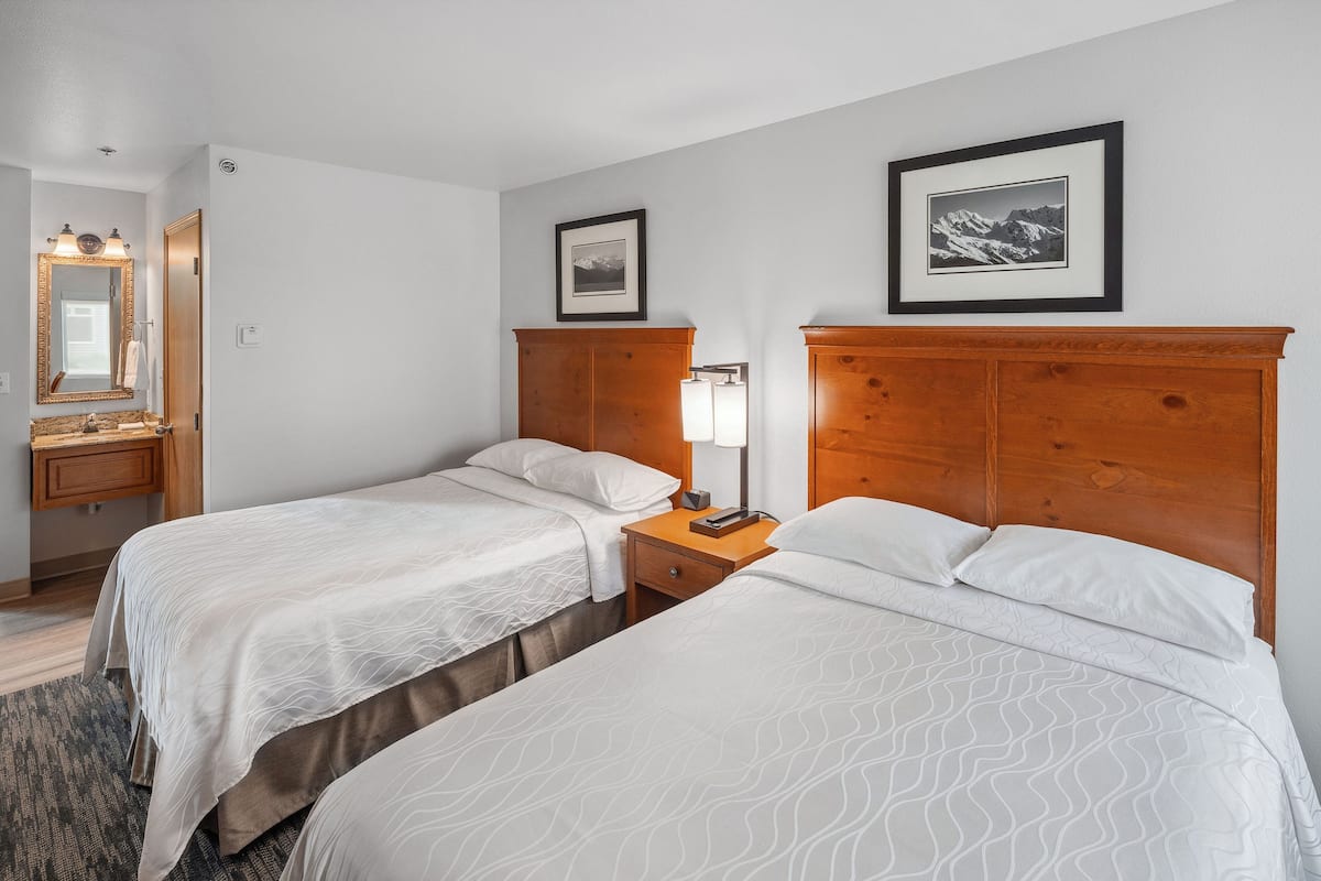 Une chambre d’hôtel avec deux lits, une tête de lit en bois, une table de chevet avec une lampe, un miroir et des tableaux encadrés accrochés au mur.