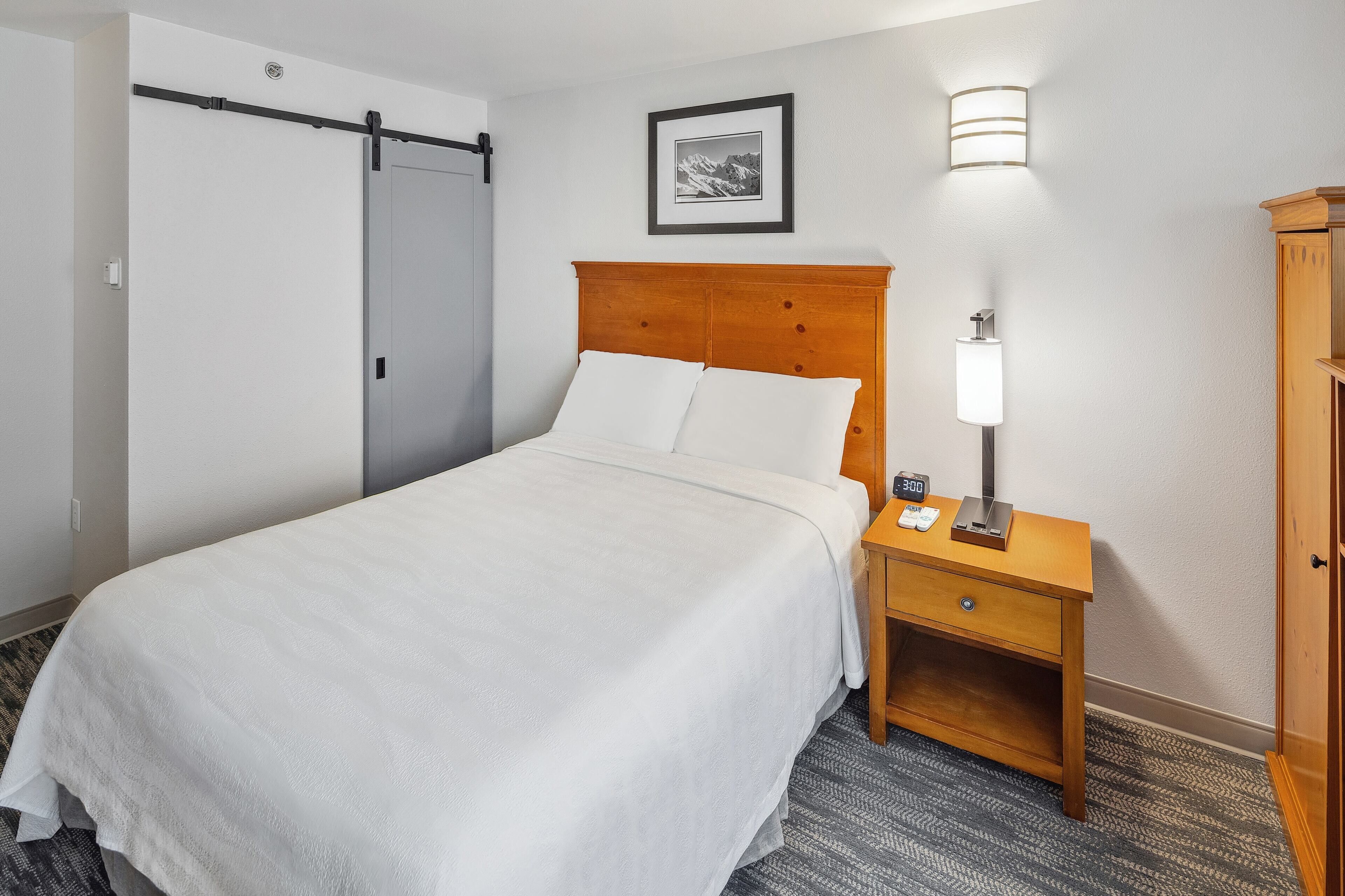 A hotel room with a bed, a nightstand with a lamp, a framed picture on the wall, and a wardrobe.