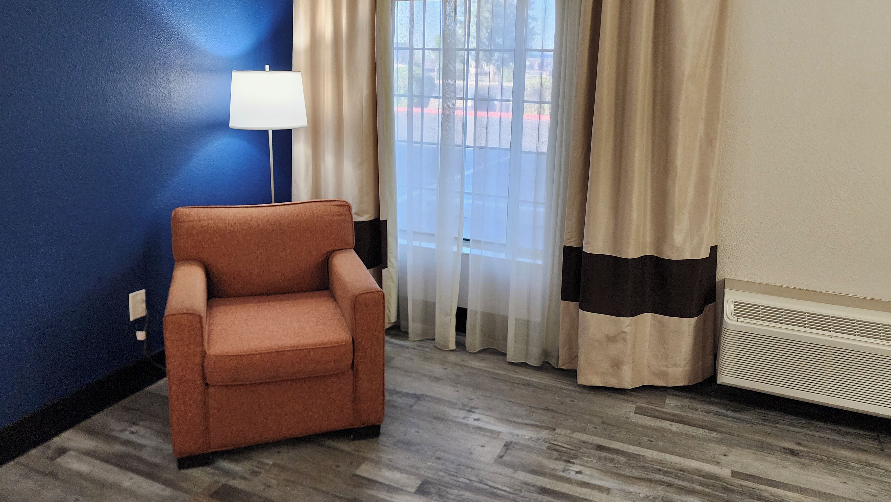 Una habitación con un sillón marrón, una lámpara blanca, cortinas color beige y una ventana que muestra una línea roja en la carretera.